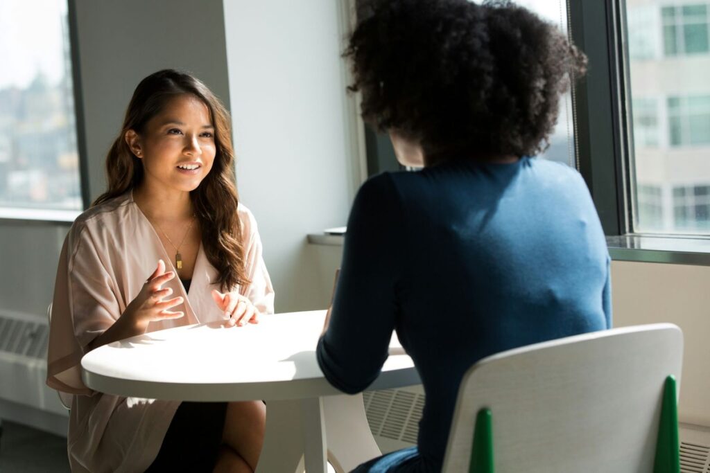 Comment Être Sûr de Soi : 15 conseils actionnables dès aujourd’hui Deux femmes qui communiquent au tour d'une table au travail